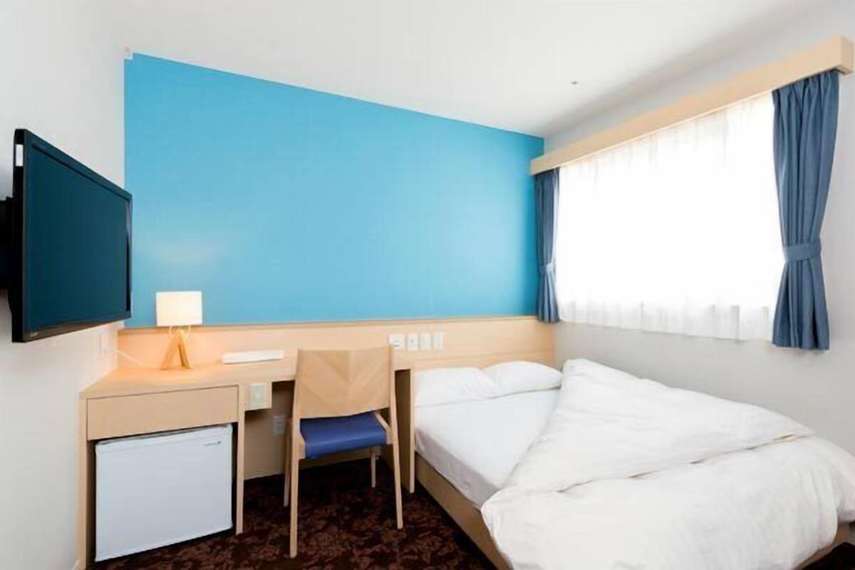 Habitación de hotel con una cama individual, un escritorio con silla, un refrigerador pequeño, un televisor montado en la pared y una ventana con cortinas.