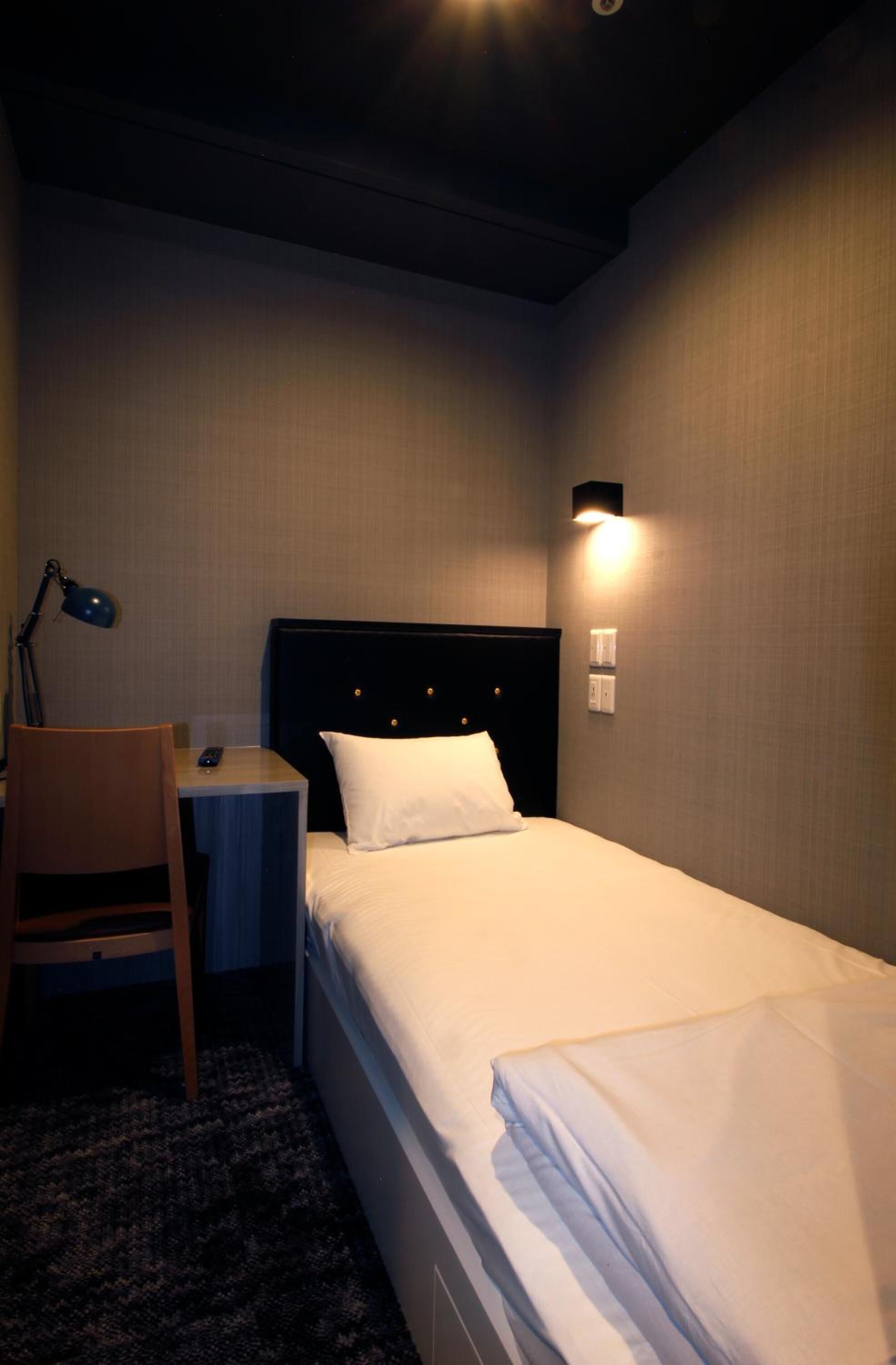 A hotel room with a single bed, a desk with a chair, a lamp, and a wall-mounted light fixture.