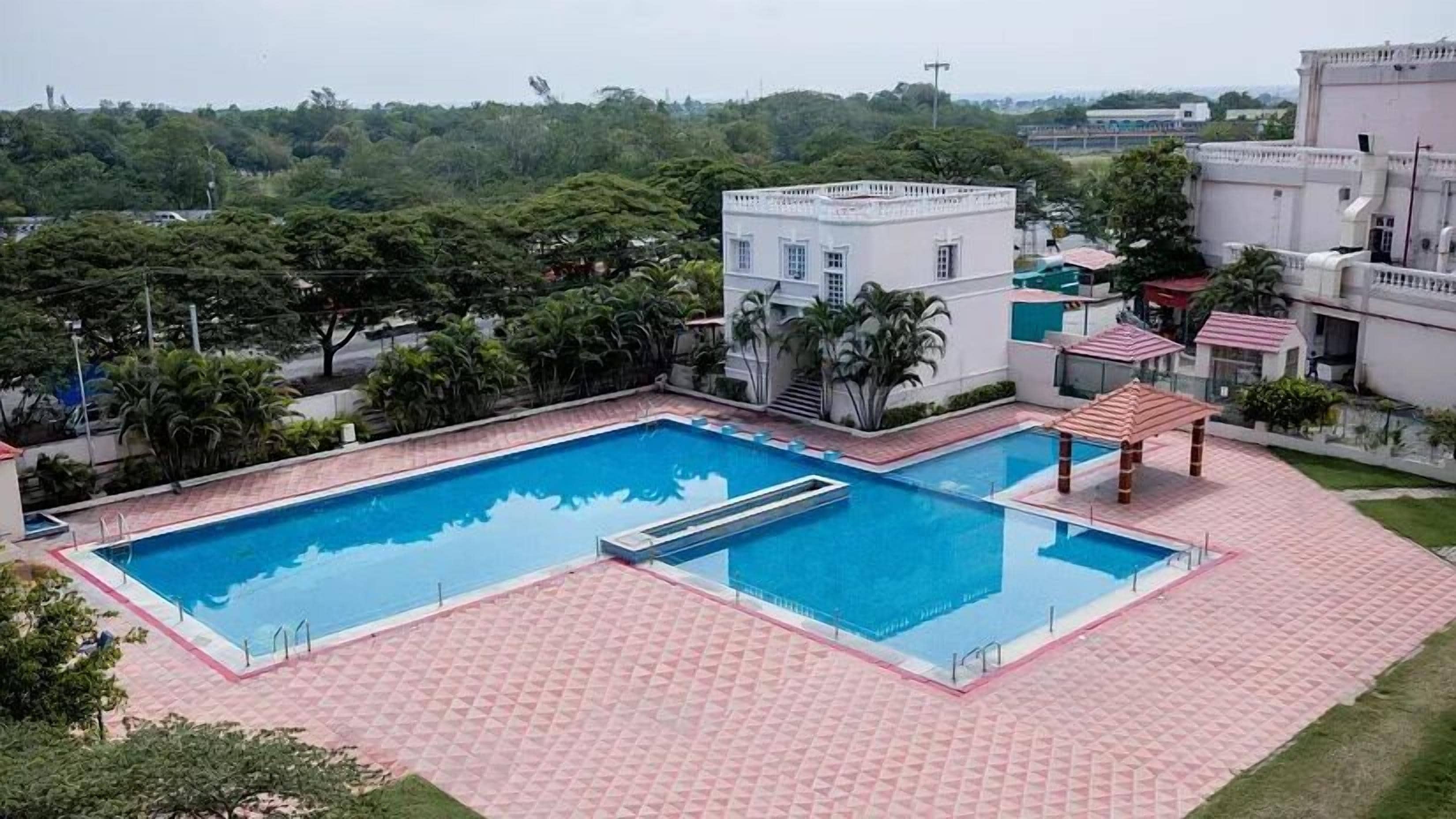 A rectangular swimming pool with a tiled deck, surrounded by greenery and buildings.