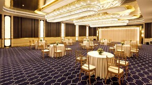 Ein geräumiger Festsaal mit runden Tischen und Stühlen, einem gemusterten Teppich und einer prunkvollen Kristallleuchter.