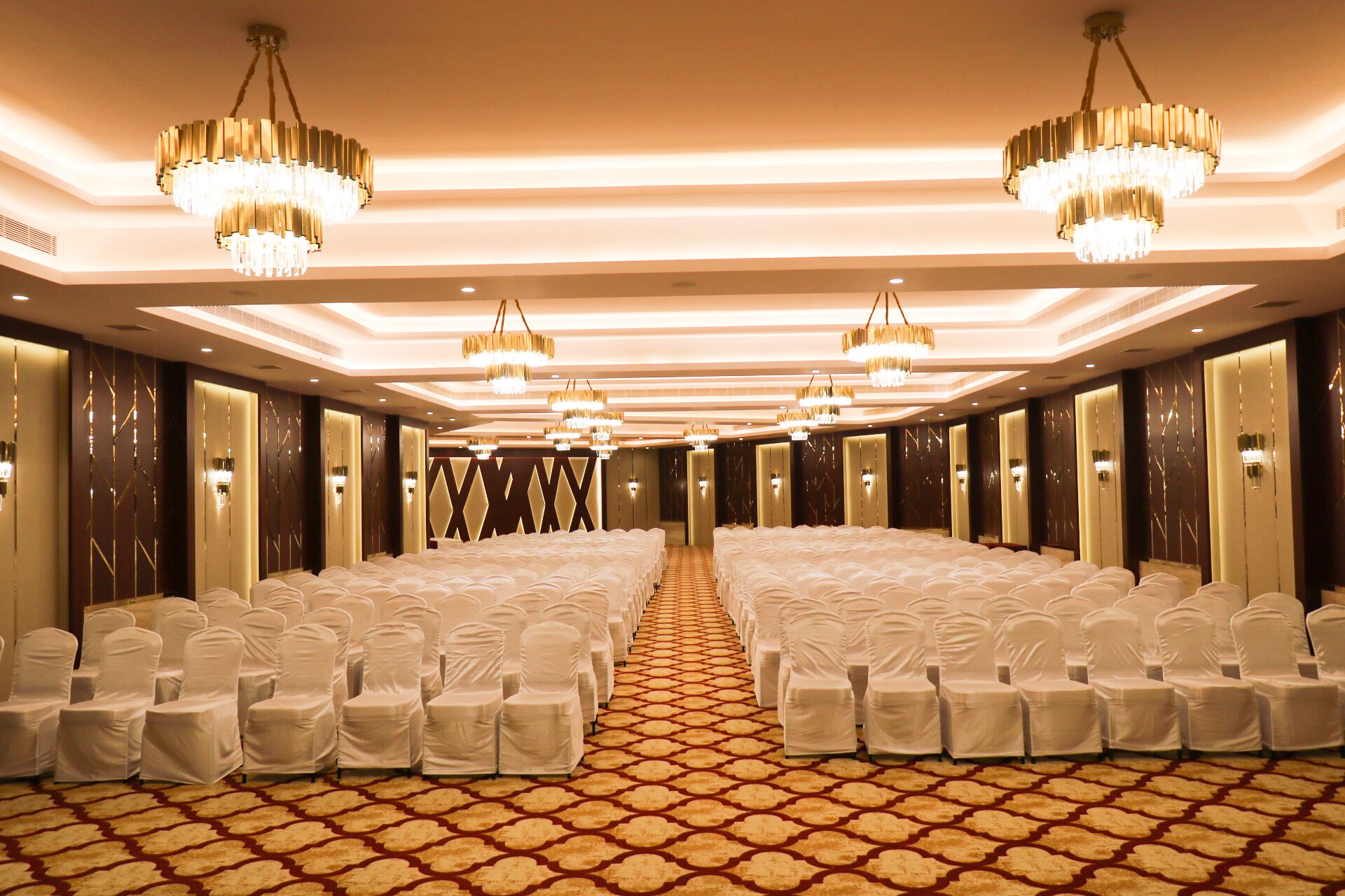 A hotel conference room with rows of white-covered chairs, chandeliers, and a patterned carpet.