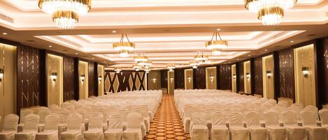 A hotel conference room with rows of white-covered chairs, chandeliers, and a patterned carpet.