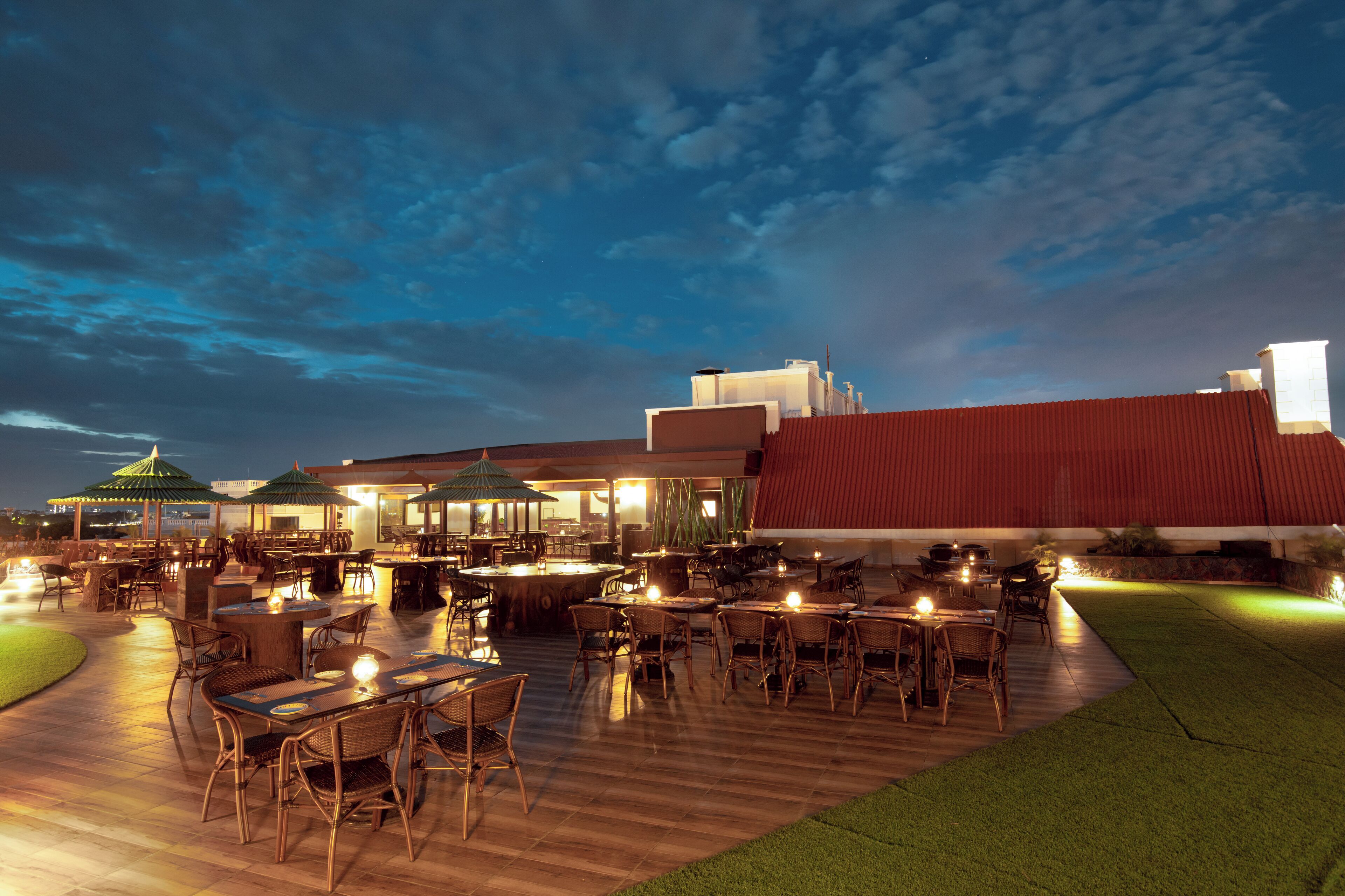 An outdoor dining area with tables and chairs, a covered seating area, and a building with a red roof in the background.