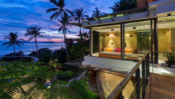 A modern house with a glass facade, a wooden deck, and a view of the ocean at dusk. - L2 Residence (Koh Samui)