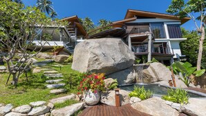 A modern house with a large rock centerpiece, a small waterfall, and a wooden deck. - L2 Residence (Koh Samui)