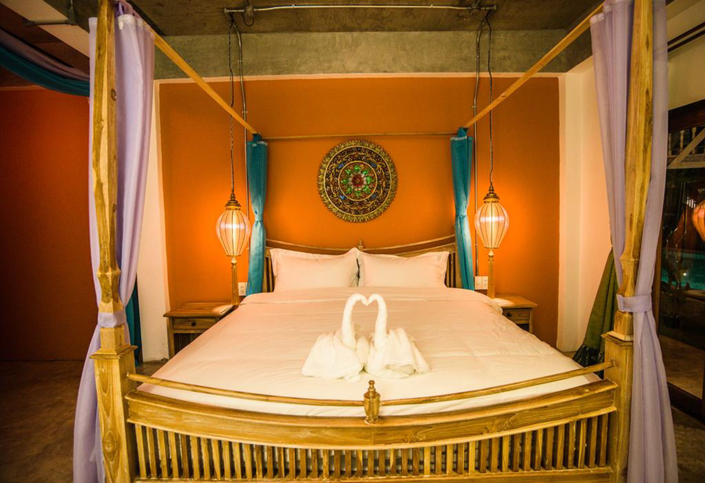 A four-poster bed with a heart-shaped towel arrangement, a decorative wall hanging, and two wall-mounted lamps.