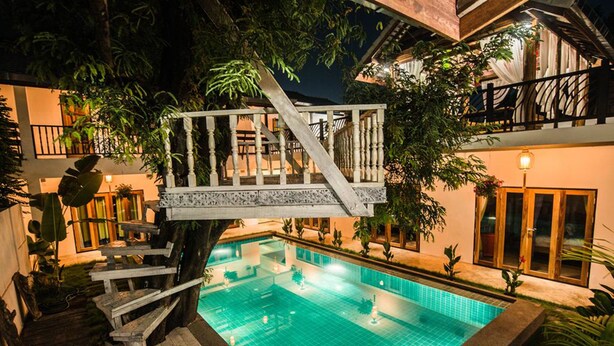 A swimming pool with a wooden deck and stairs, surrounded by lush greenery and a building with balconies. - Garden Yard Inn Chiangmai (Chiang Mai)