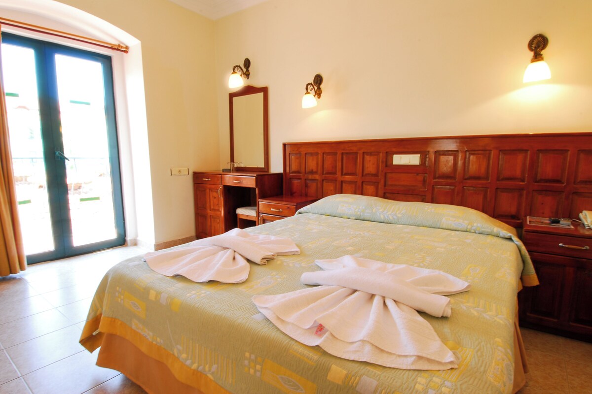 Habitació d'hotel amb un llit gran, un panell de fusta al capçal, una taula de nit, un aparador i una porta que dona a una zona exterior.