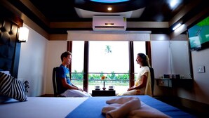 A hotel room with a bed, a bedside table with a vase, a television, and a window with a view of greenery.