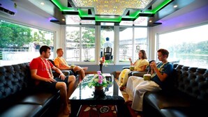 A group of people seated on a black sofa in a room with a glass table, a decorative ceiling, and a view of a body of water.