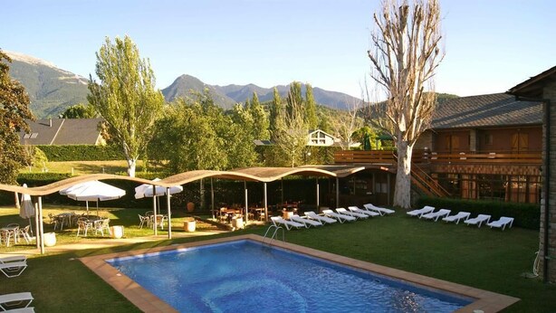 Piscina con sillas de descanso y sombrillas, rodeada de un césped cuidado y con montañas al fondo.