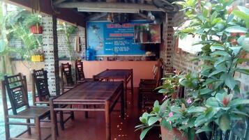 A covered outdoor seating area with wooden tables and chairs, a blue signboard, and potted plants.
