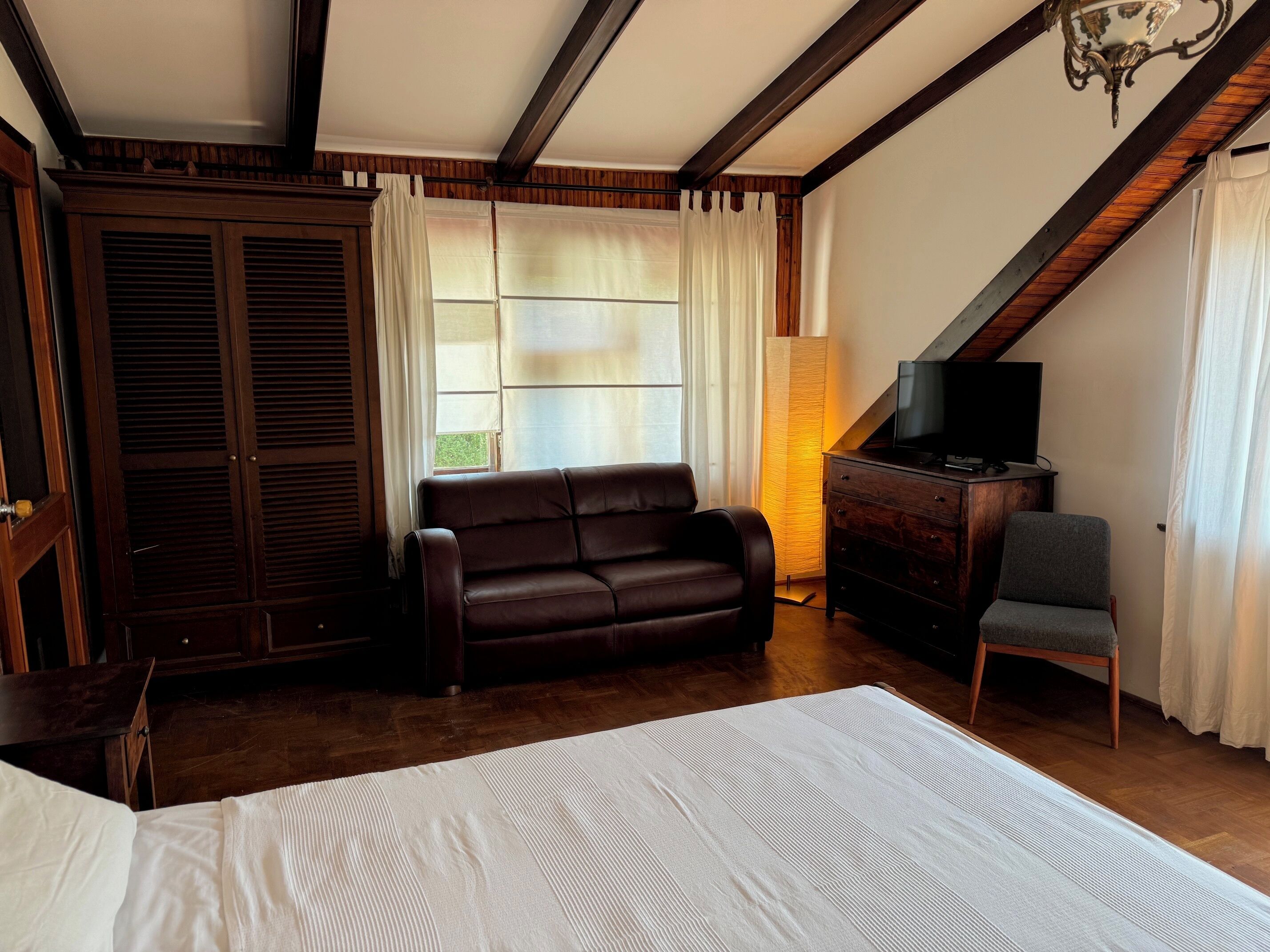 A bedroom with a bed, a dark brown leather sofa, a wooden dresser with a television, a chair, and a window with curtains.
