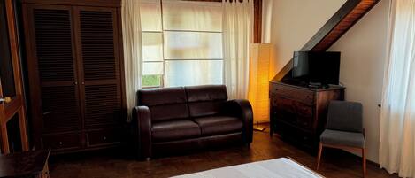 Ein Schlafzimmer mit einem Bett, einem dunkelbraunen Ledersofa, einem Holzschrank mit Fernseher, einem Sessel und einem Fenster mit Vorhängen.