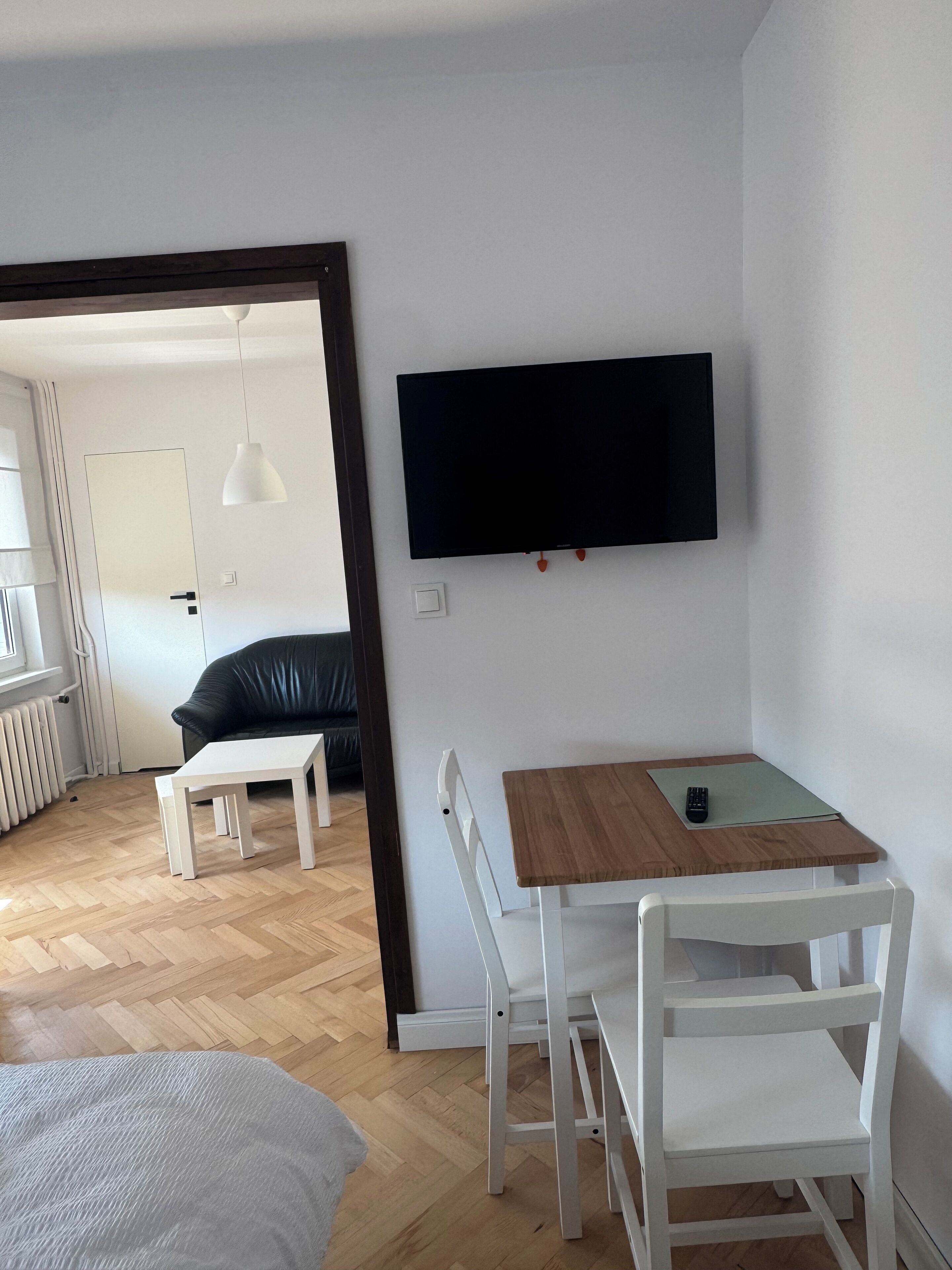 A modern living room with a flat-screen TV mounted on the wall, a wooden dining table with chairs, a white sofa, and a small white table.
