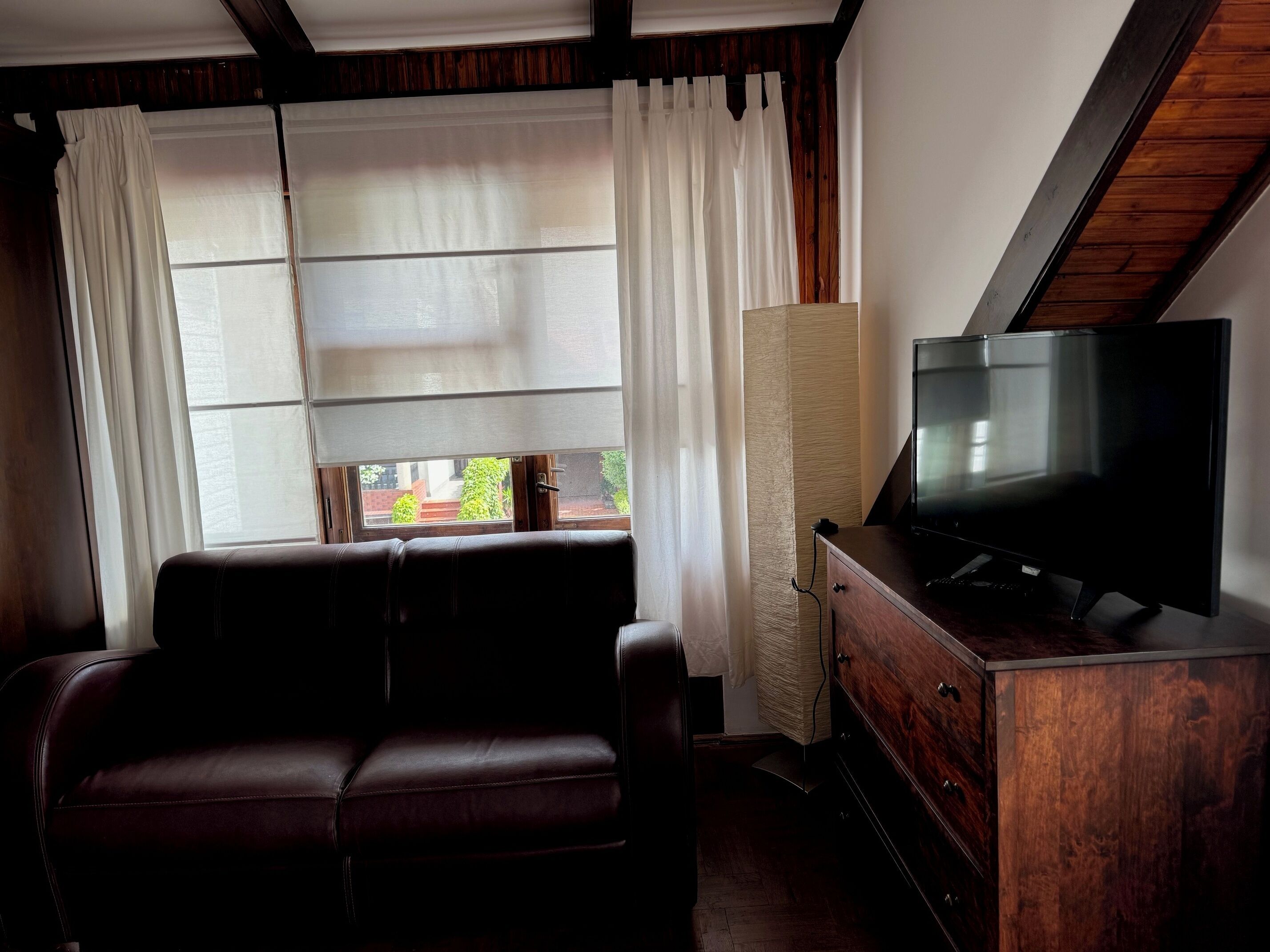 Una habitación con un sofá de cuero, un televisor sobre un mueble de madera y una ventana con cortinas blancas.