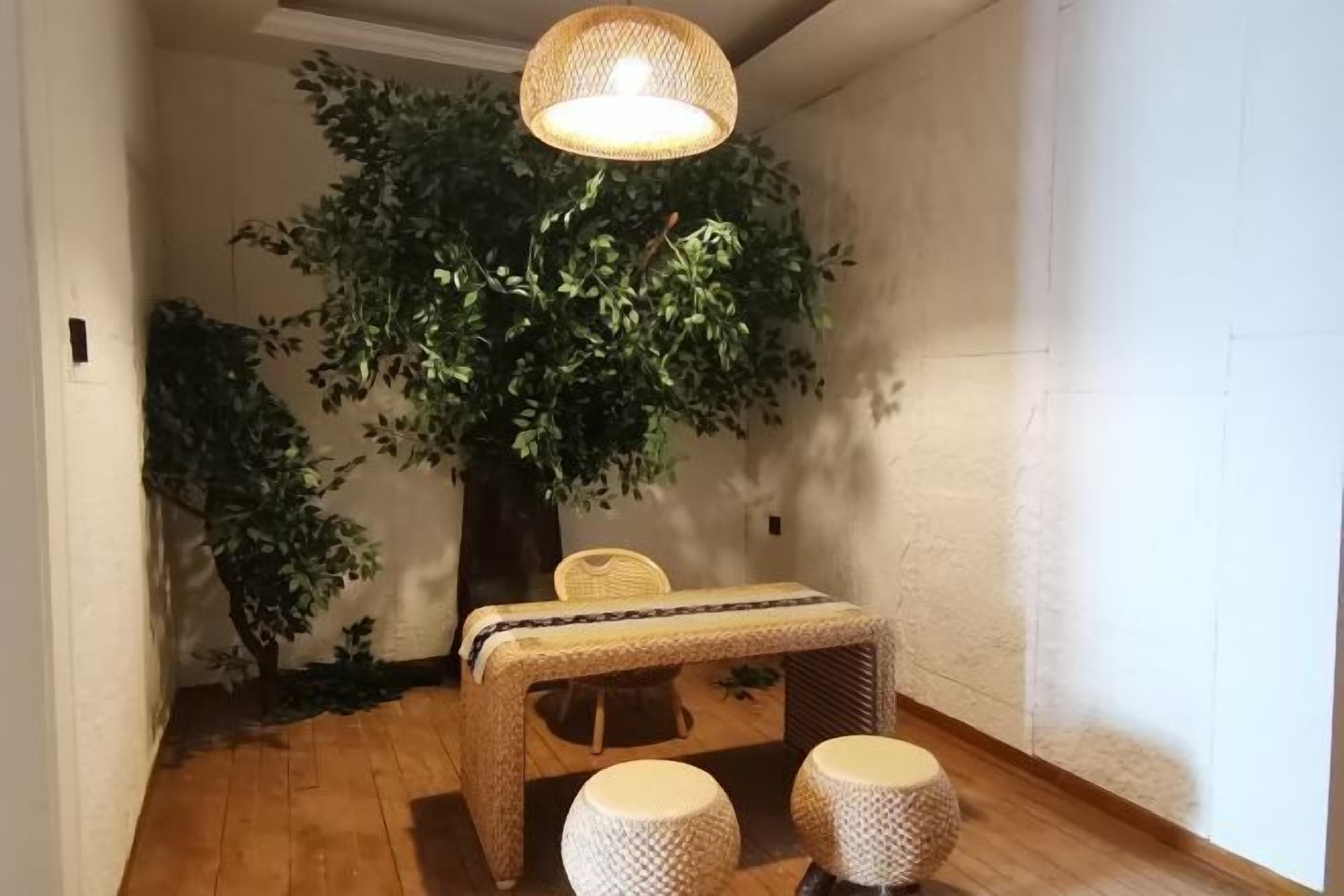 Una habitación con piso de madera, una pequeña mesa con mantel tejido, dos taburetes redondos, una silla de mimbre y un árbol artificial grande.