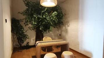 Una habitación con piso de madera, una pequeña mesa con mantel tejido, dos taburetes redondos, una silla de mimbre y un árbol artificial grande.