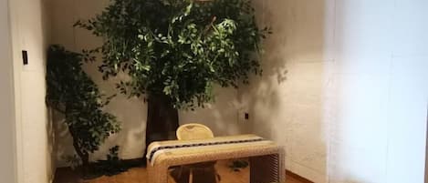 A room with a wooden floor, a small table with a woven tablecloth, two round stools, a wicker chair, and a large artificial tree.