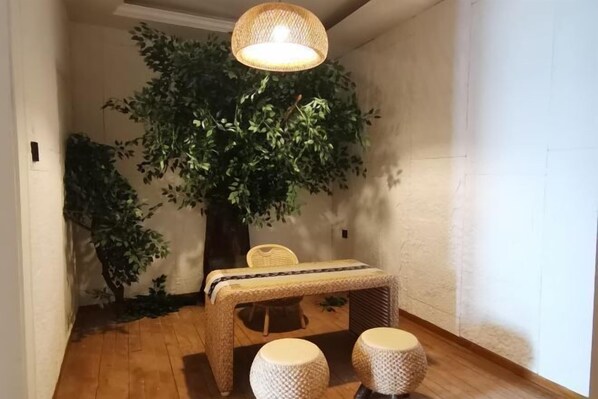 Een kamer met een houten vloer, een tafeltje met een geweven tafelkleed, twee ronde krukjes, een rieten stoel en een grote kunstboom.