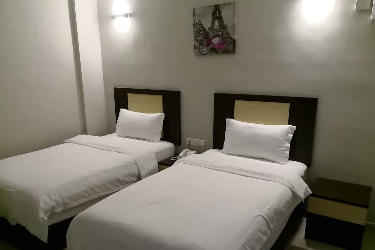 A hotel room with two single beds, each with white bedding and headboards. There is a small bedside table with a phone on it, and a wall-mounted light fixture above the beds.
