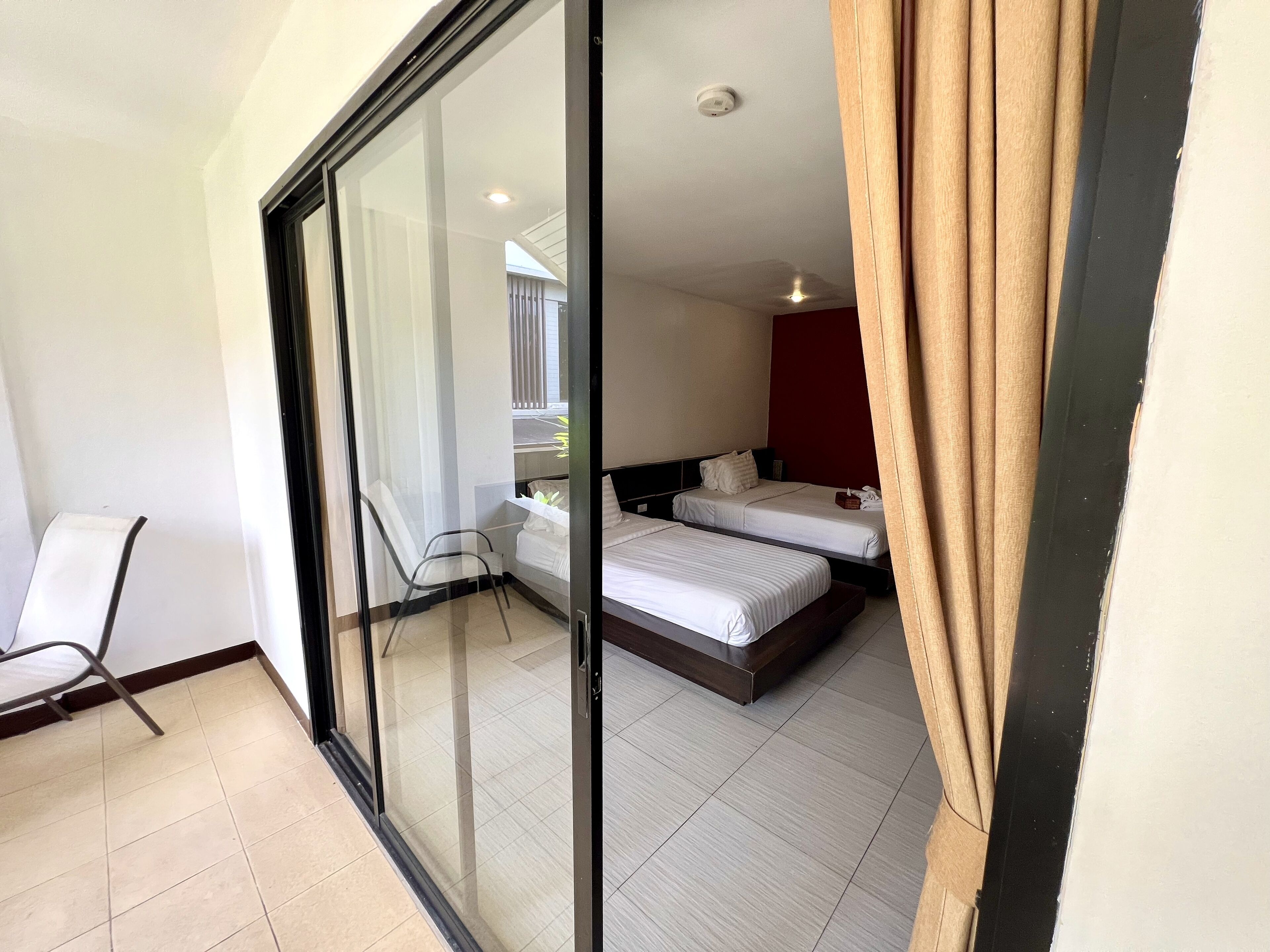 Une chambre d’hôtel avec une porte vitrée donnant sur un balcon, deux lits, une chaise et un mur d’accent de couleur rouge.