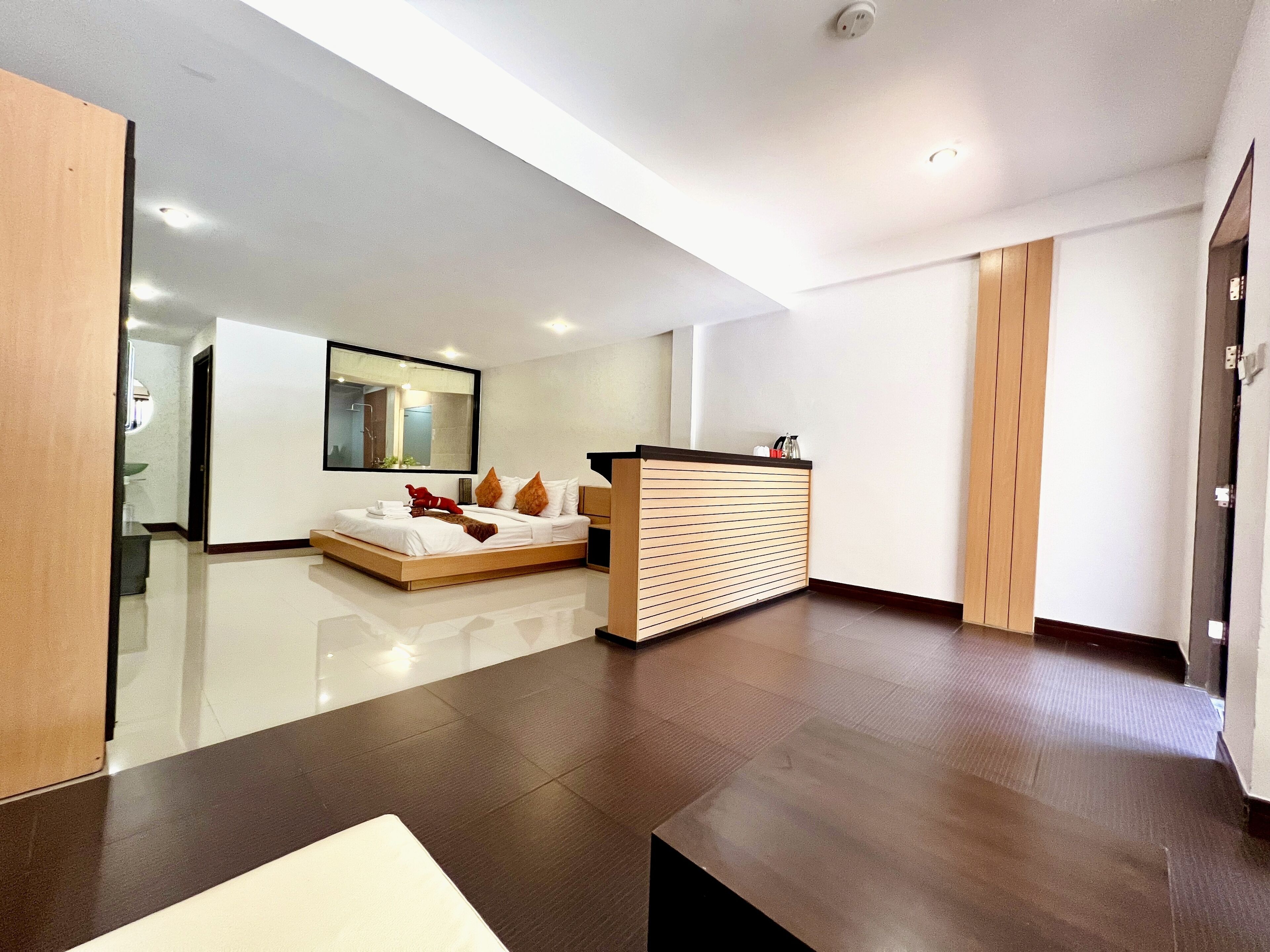 A modern hotel room with a large bed, a wooden side table, a mirror, and a small kitchenette area.