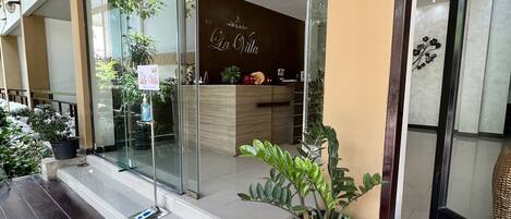 A hotel entrance with a 'WELCOME' mat, glass doors, a reception desk, and potted plants.