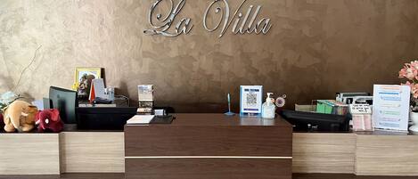 A reception desk at La Villa Hotel with a wooden finish, a framed photo, a small statue, a stack of brochures, a QR code sign, and a water bottle.