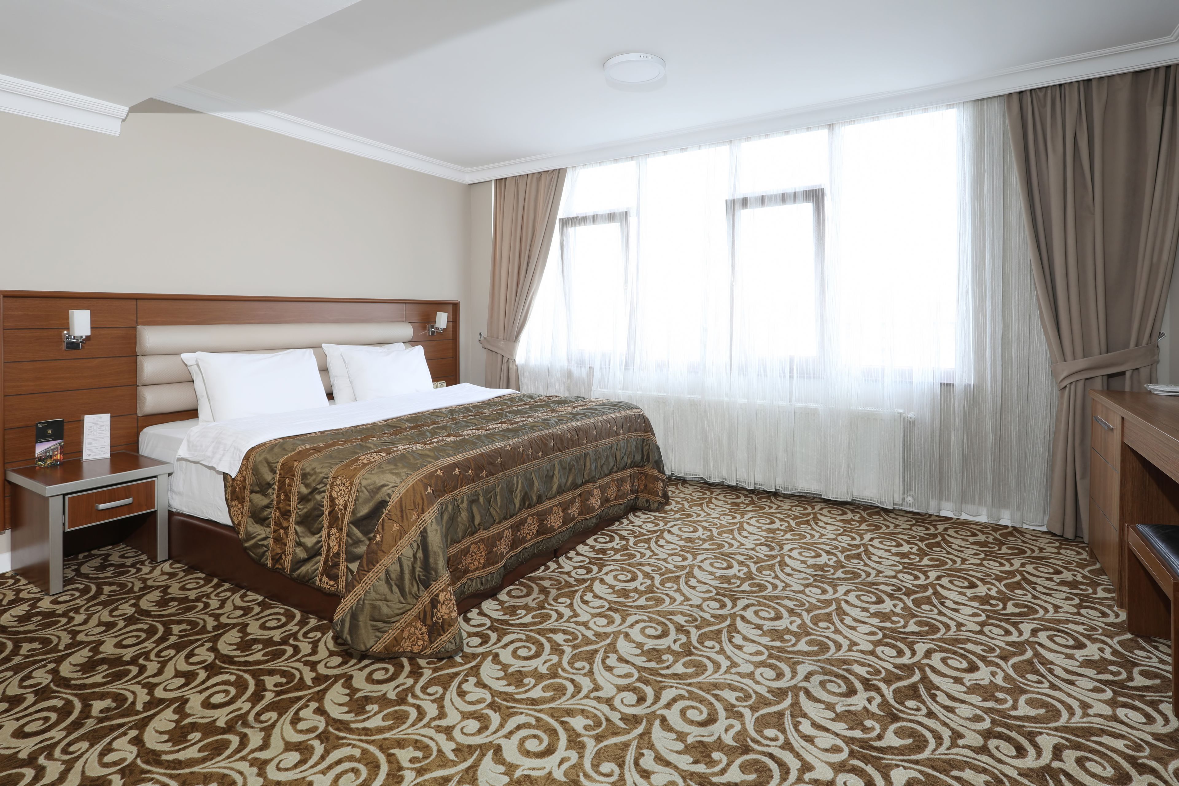 A hotel room with a large bed, a nightstand, a wooden headboard, a patterned carpet, and a large window with curtains.