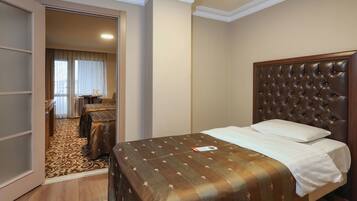 Habitación de hotel con una cama grande, suelo de madera, alfombra estampada, armario y vistas a otra habitación con escritorio y silla.