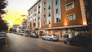 Un hotel de varios pisos con fachada moderna, que destaca un letrero de 'AVIS', una tienda Vodafone y una tienda 'SEMT BÜFE'.