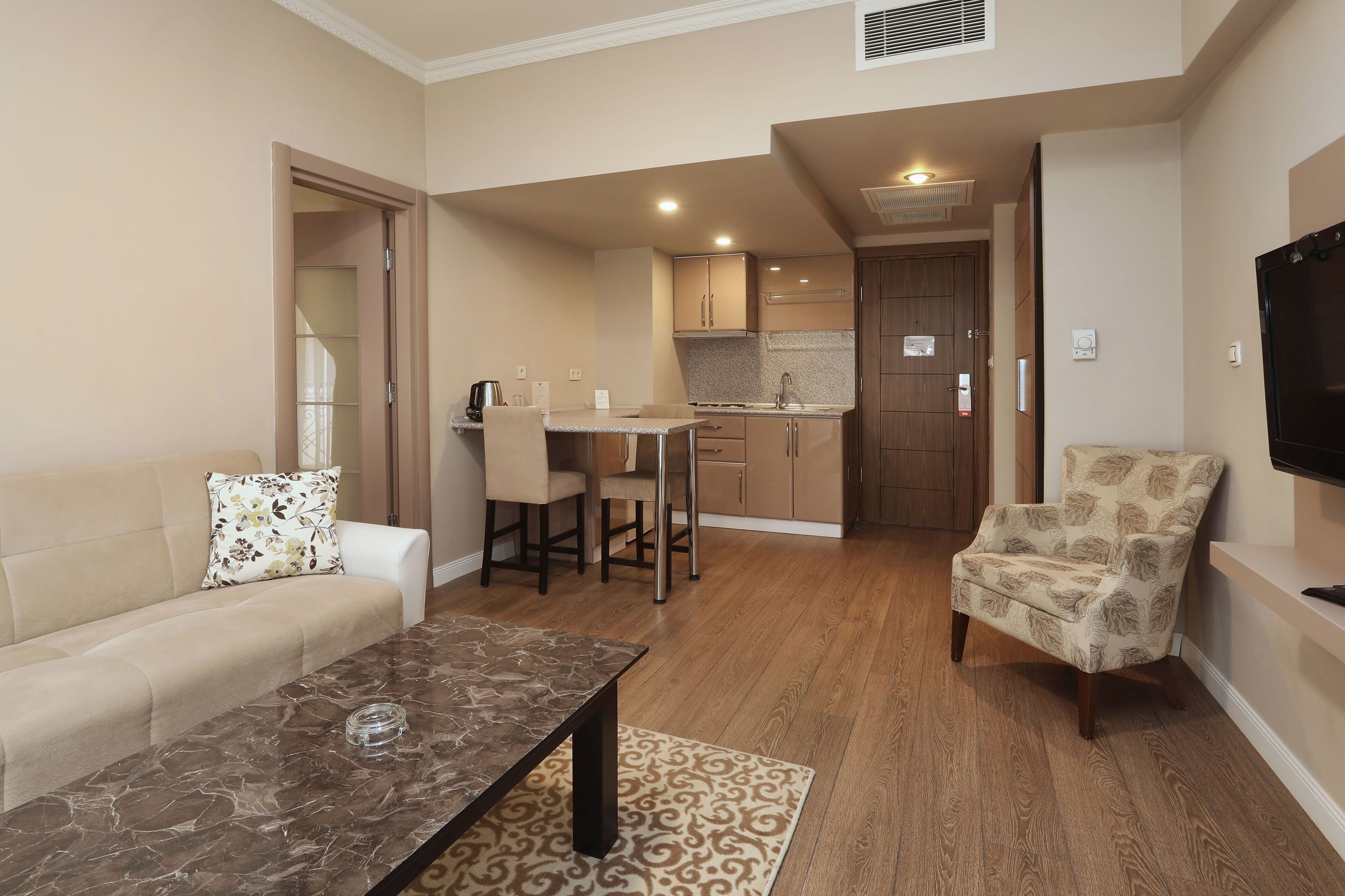 A living room with a sofa, a coffee table, and a patterned armchair. There is a kitchen area with a dining table and chairs.