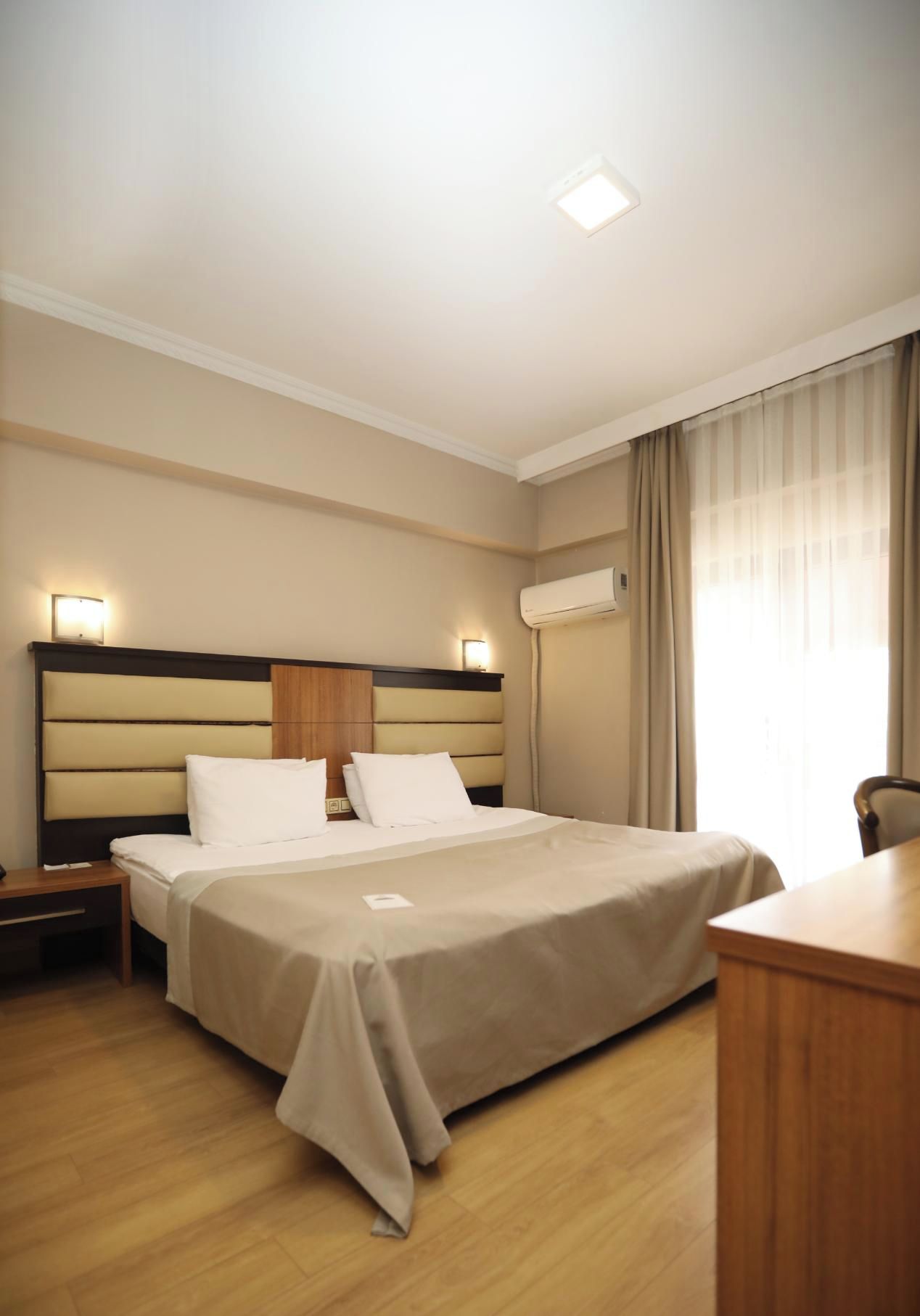 A hotel room with a bed, bedside tables, a desk, and an air conditioning unit.
