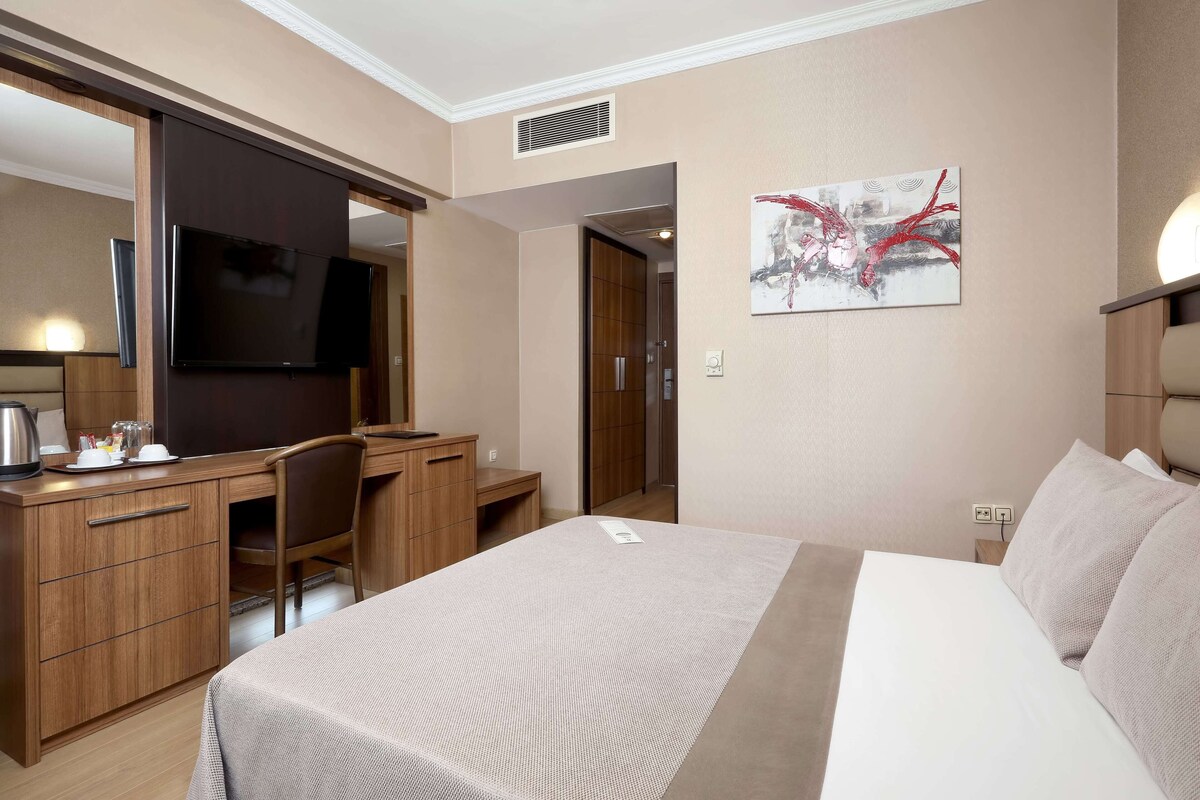 A hotel room with a bed, a desk with a chair, a television, and a painting on the wall.