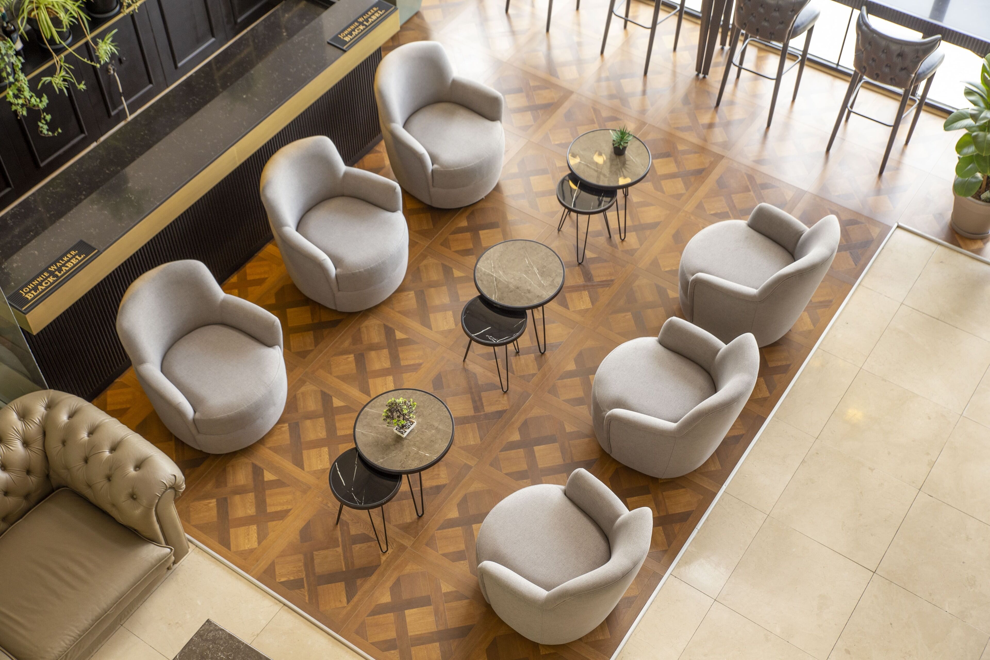 Un vestíbulo moderno con un suelo de estampado geométrico, varios sillones grises y pequeñas mesas redondas con plantas en macetas.