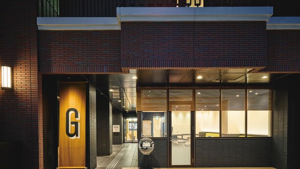 Een gebouwtoegang met een groot, verlicht 'G'-bord, een glazen deur en een gevel van baksteen.