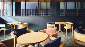 Een modern interieur met ronde houten tafels en geweven stoelen, een donkere muur en uitzicht op een gebouw door grote ramen.