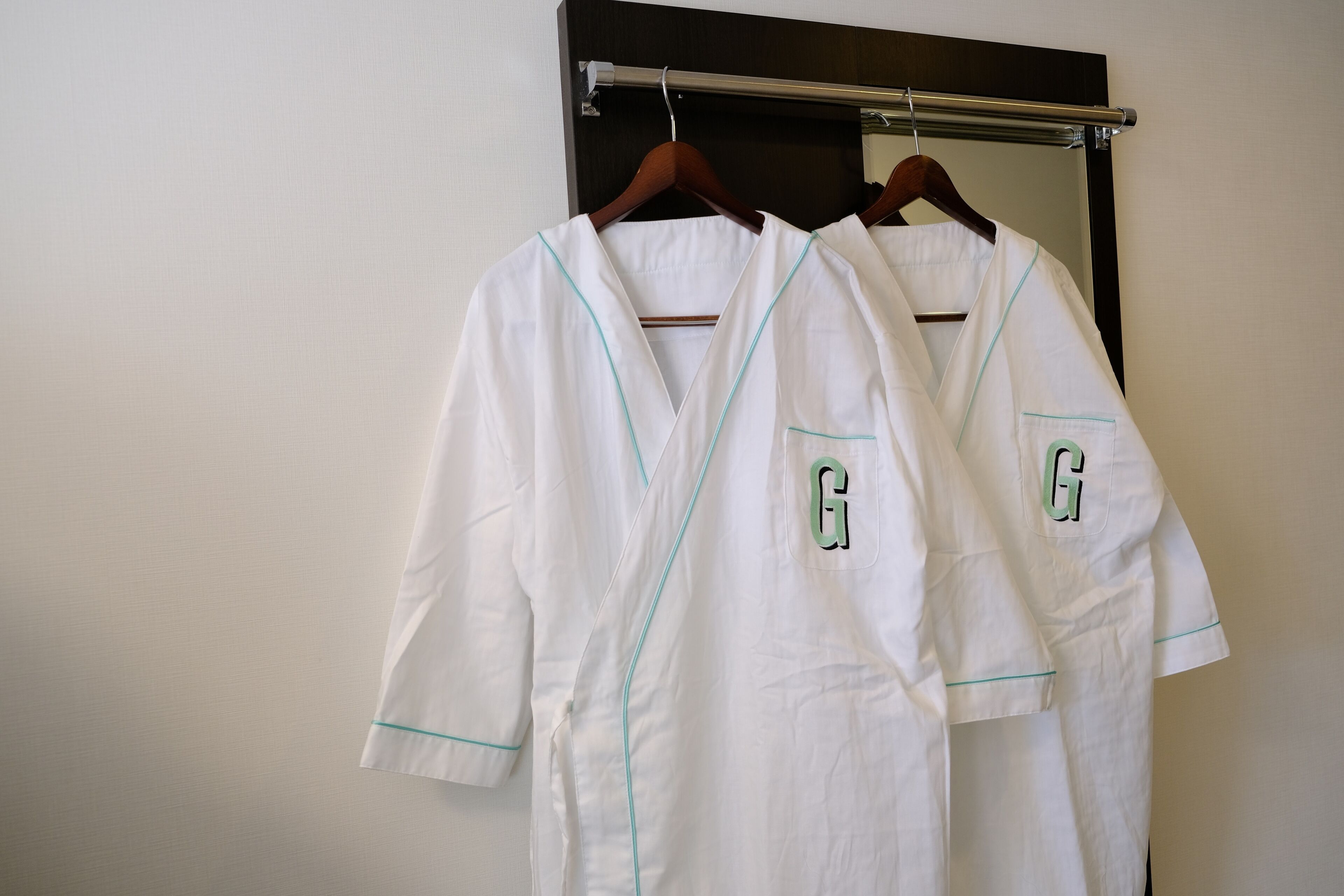 Two white robes with teal piping and the letter 'G' on a hanger.