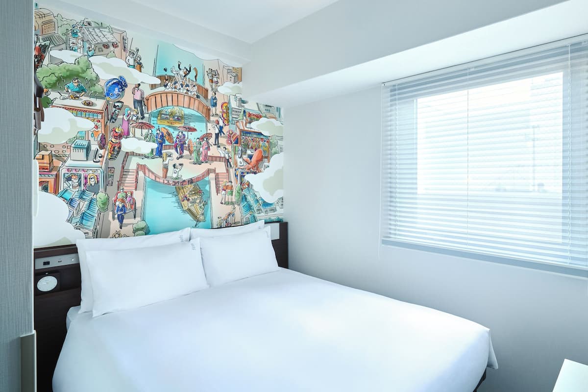 Een hotelkamer met een groot bed, een raam met zonwering en een kleurrijke muurschildering.