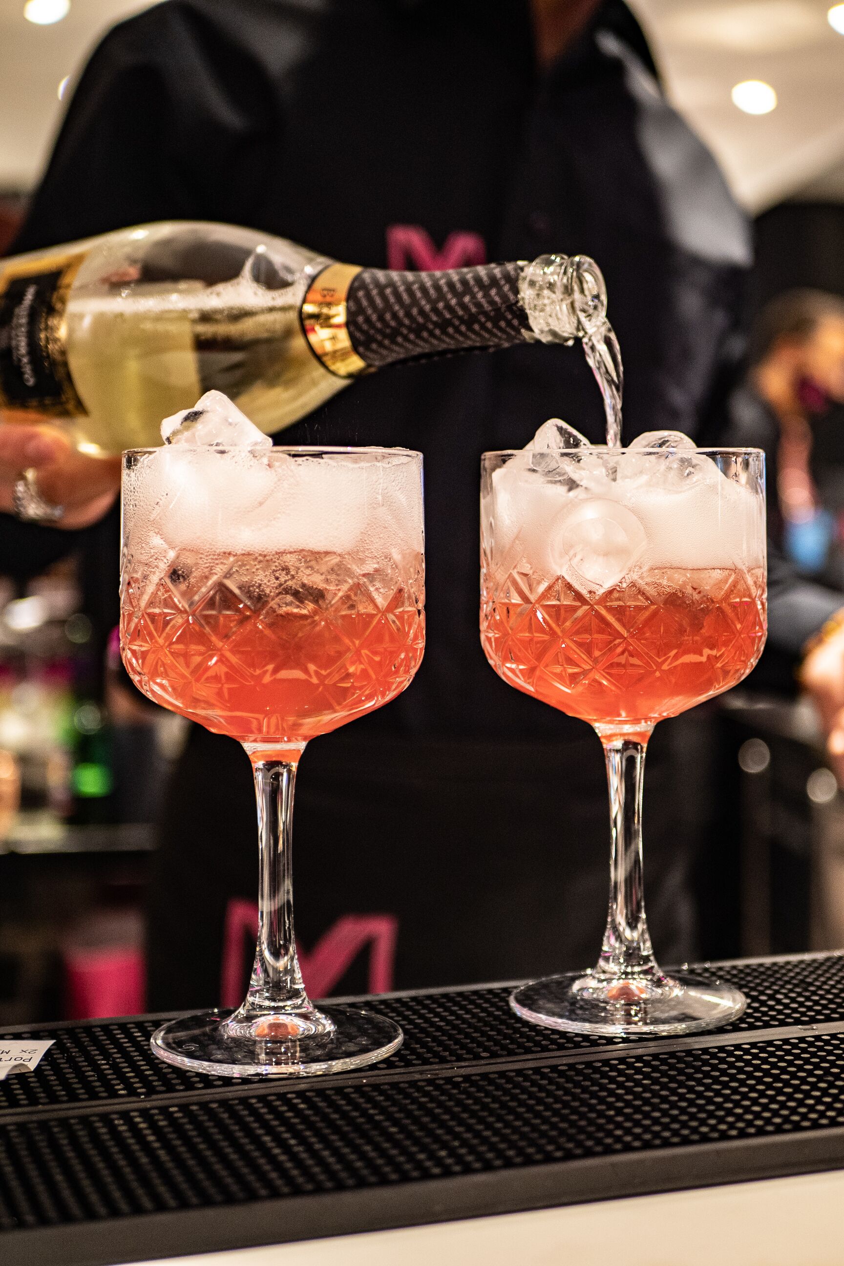 Two glasses of a red cocktail with ice cubes, being filled with a clear liquid from a bottle.