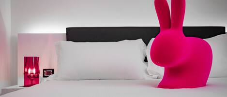 Una cama con sábanas blancas, cabecero negro y un objeto rosa grande con forma de conejo.