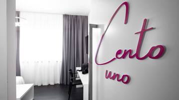 A modern hotel room with a bed, a desk, and a chair. The room has a large window with curtains. There is a sign on the wall that reads "Cento uno".