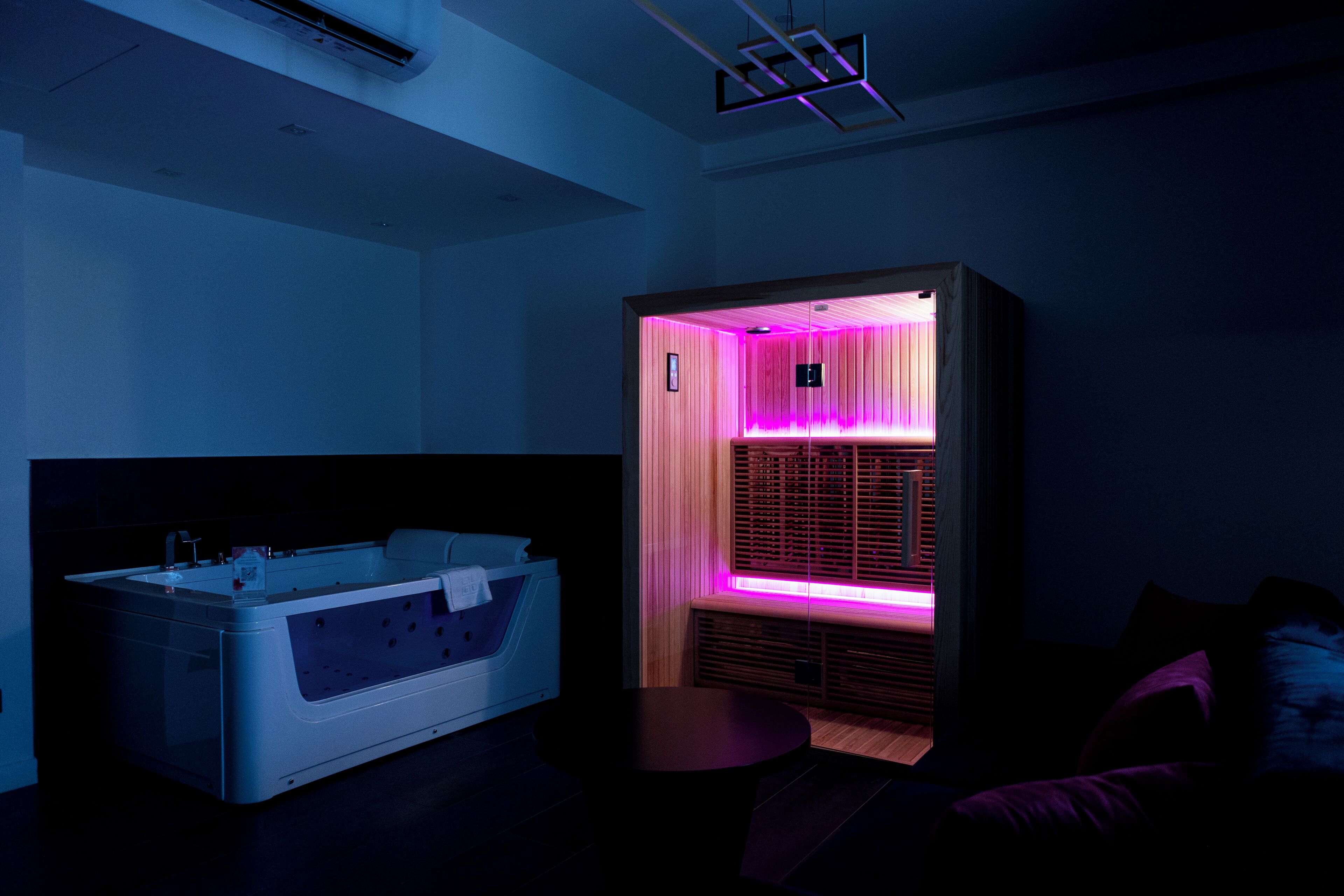 Una moderna sala spa con una vasca da bagno ampia, un lettino da massaggio e un mobile in legno illuminato da una luce rosa.