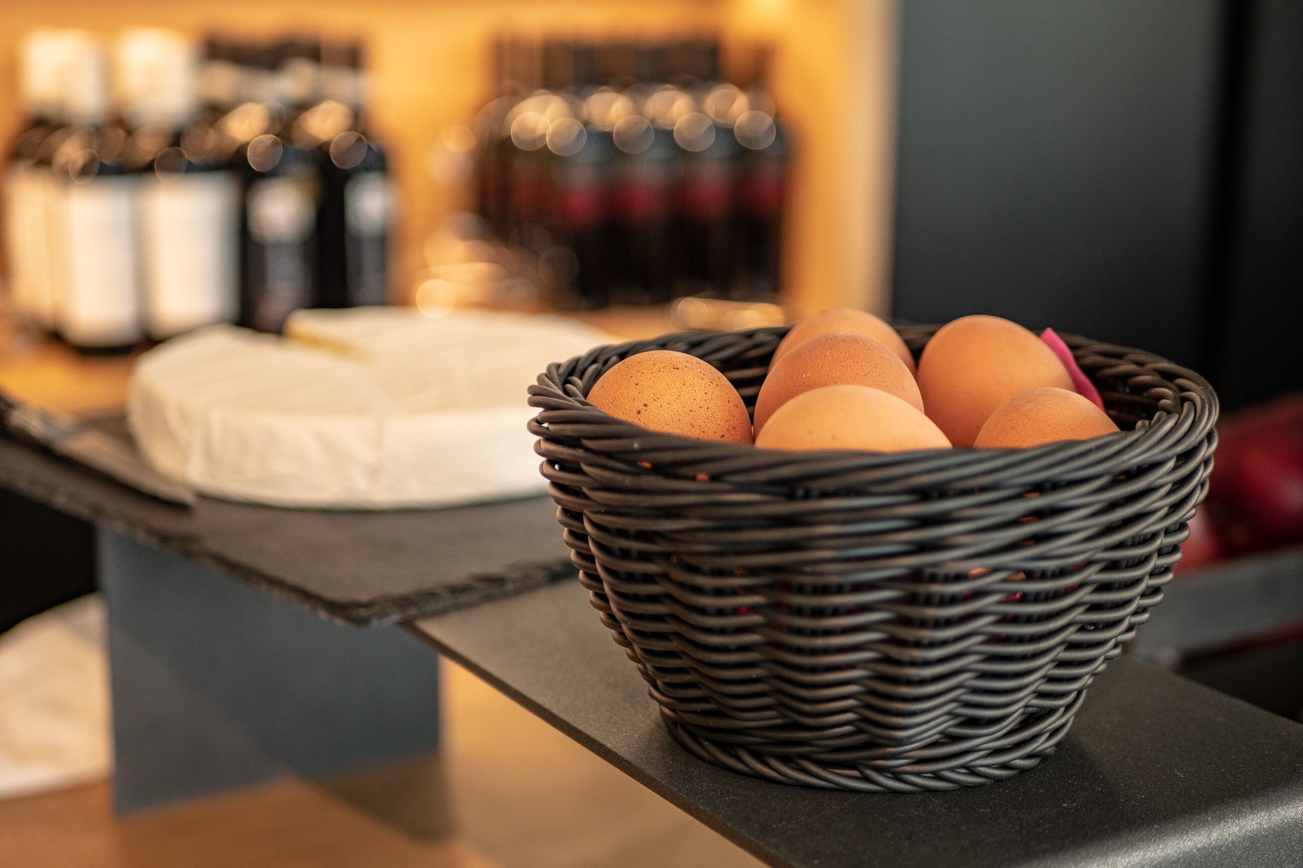 A wicker basket with eggs on a counter, with bottles and a cake in the background.