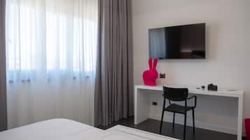 Quarto de hotel com uma cama, uma escrivaninha com cadeira, uma televisão na parede e uma janela com cortinas.