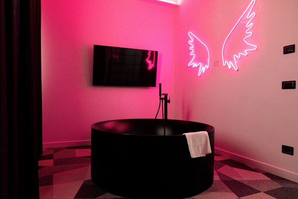 Une salle de bain moderne avec une baignoire noire, un éclairage néon en forme d’aile et un téléviseur fixé au mur.