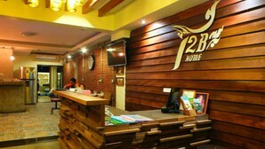 A reception desk with a wooden finish, a clock, and a television. There is a counter with a stack of books and a framed picture. The wall has a sign with the text "12B HOME". - T2B Home - Adults Only - Hostel (Chiang Mai)