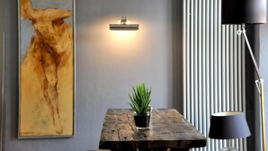 Una habitación con un cuadro abstracto grande, una planta en maceta sobre una mesa de madera, una lámpara de pared, un radiador y una lámpara de pie.
