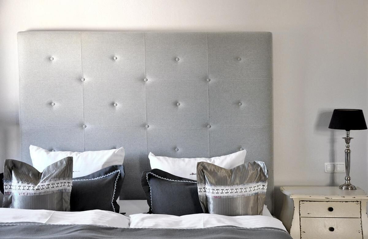 Een net opgemaakt bed met een grijze gestuufde hoofdbord, witte en grijze kussens en een nachtkastje met een lamp.